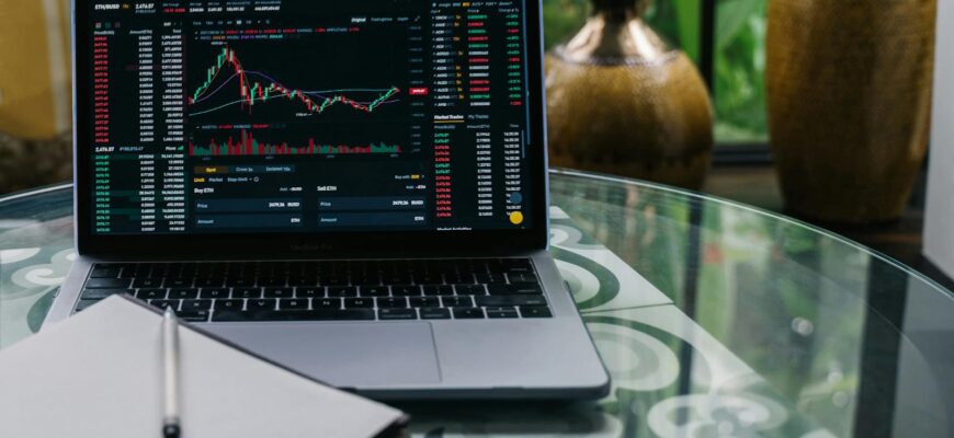 Laptop displaying cryptocurrency stocks and graphs on a glass table with a notepad.