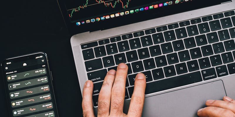 Hands typing on a laptop with cryptocurrency charts and smartphone display in view, focusing on digital finance.